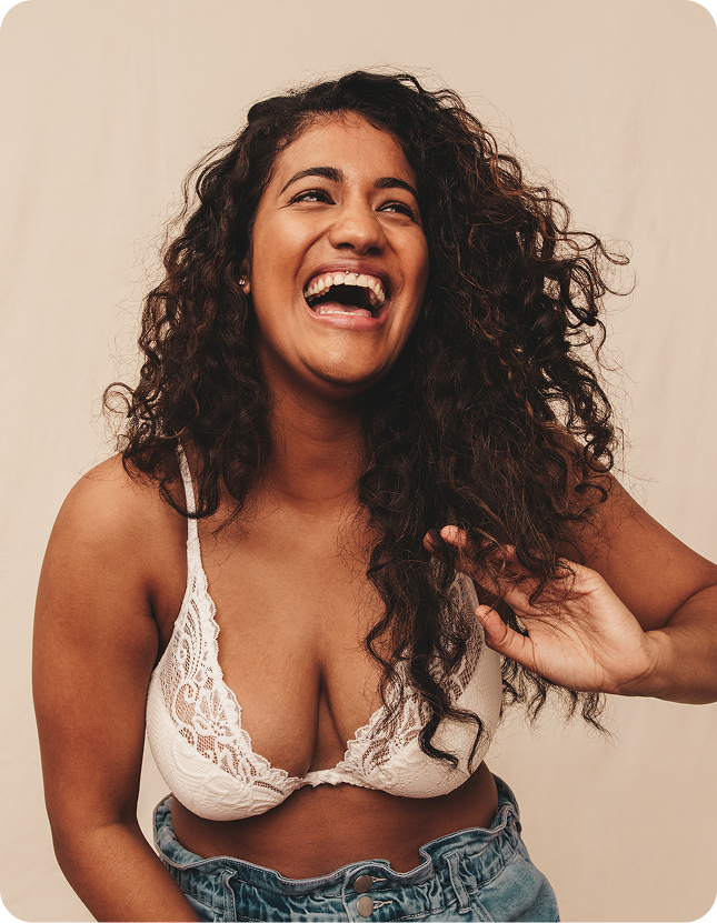 Woman laughing with curly hair wearing a white lace top against a beige background